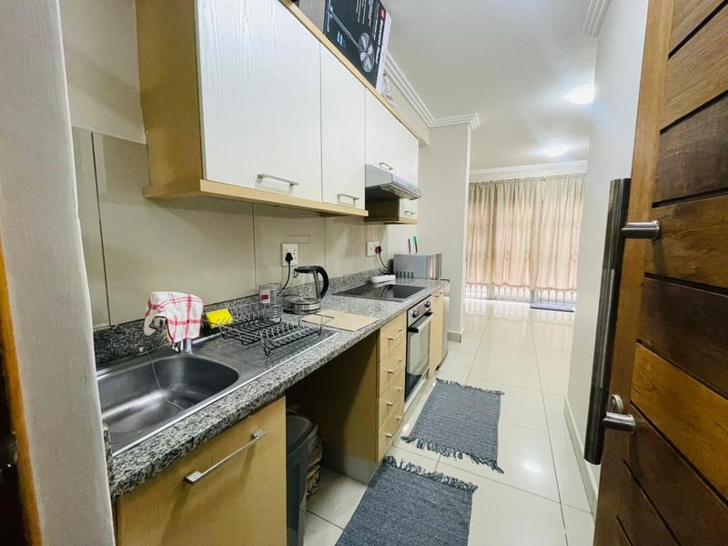 A functional galley-style kitchen featuring granite countertops, built-in cabinetry, integrated oven, and hob, leading into an open-plan living area with tiled floors.