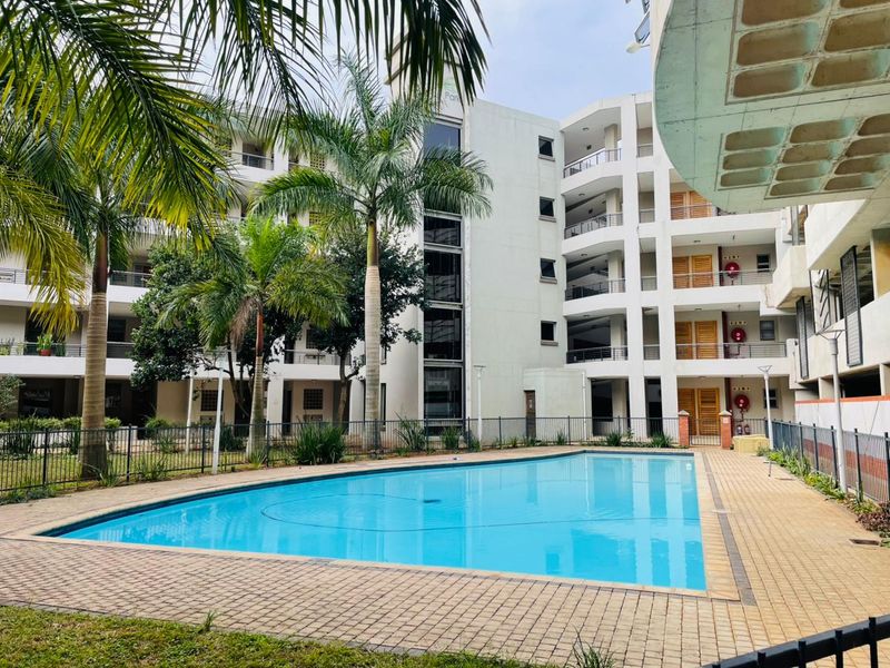 A well-maintained communal swimming pool area featuring paved decking, metal fencing, and views of the multi-story apartment building facade.