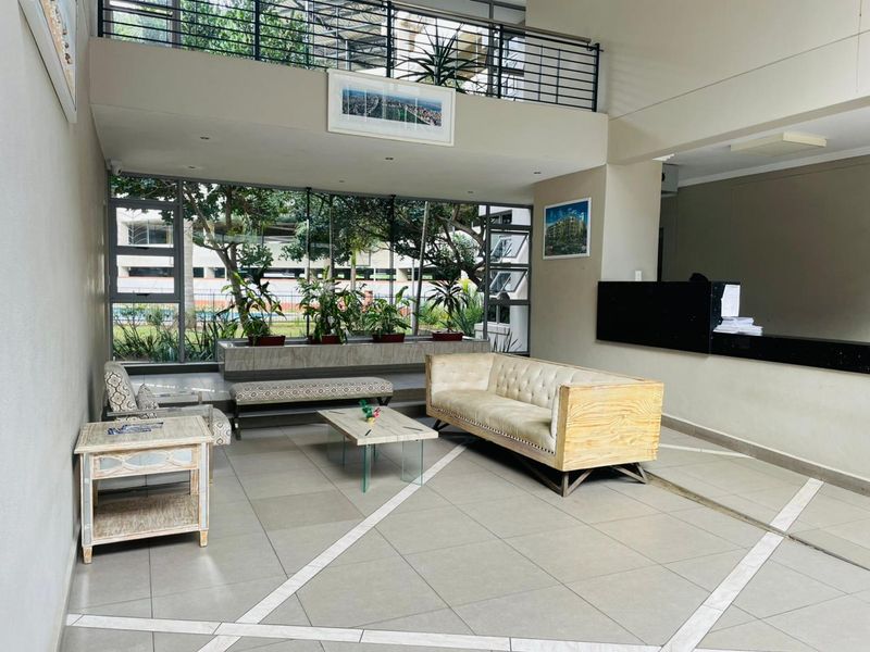Spacious apartment building entrance lobby featuring a reception desk, mezzanine level with metal balustrade, and large windows overlooking the exterior grounds.