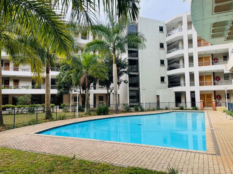 A well-maintained communal swimming pool area set within a modern apartment complex, featuring paved surrounds and landscaped garden elements.