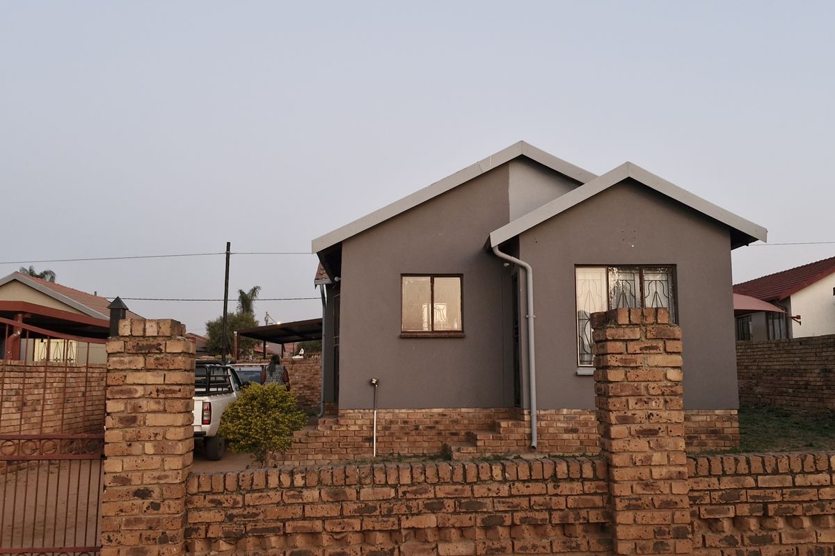 Front view of a single-story residential house featuring a grey facade, brick boundary wall, and secure gated entrance.