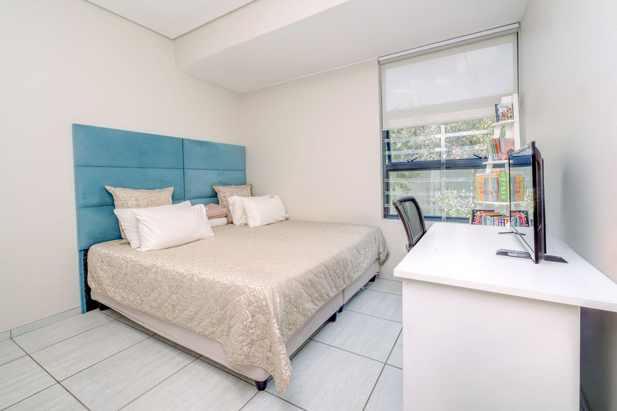 A well-maintained bedroom featuring neutral tiled flooring, clean white walls, and a large window with a roller blind for natural light.