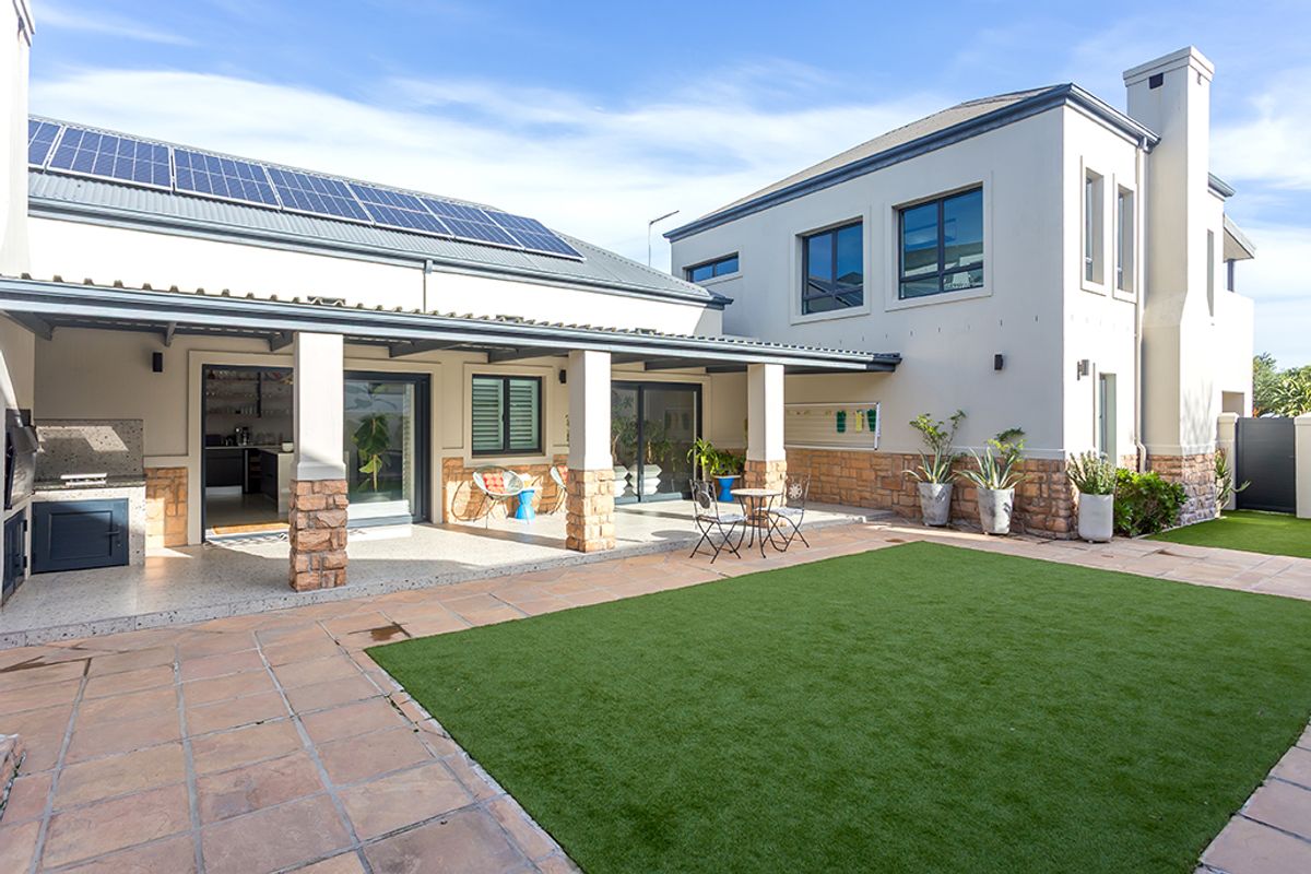 Spacious rear exterior featuring a covered patio with built-in braai, stone-clad pillars, solar panels, and a well-maintained artificial lawn area.