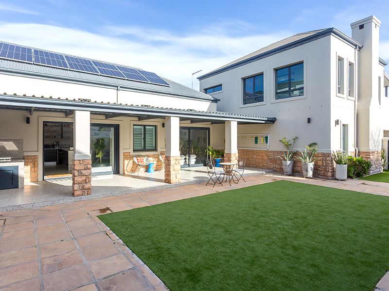 Spacious rear exterior featuring a covered patio with built-in braai, stone-clad pillars, solar panels, and a well-maintained artificial lawn area.