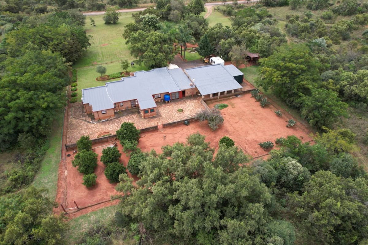 Aerial view of a brick farmhouse featuring a metal roof, expansive grounds, mature trees, and a paved patio area in a rural setting.