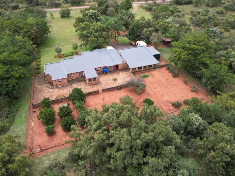 Aerial view of a brick farmhouse featuring a metal roof, expansive grounds, mature trees, and a paved patio area in a rural setting.