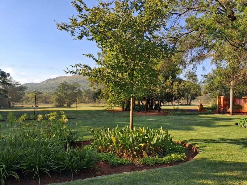 A well-maintained, expansive garden area featuring lush green lawns, established trees, and landscaped flower beds with scenic mountain views in the background.
