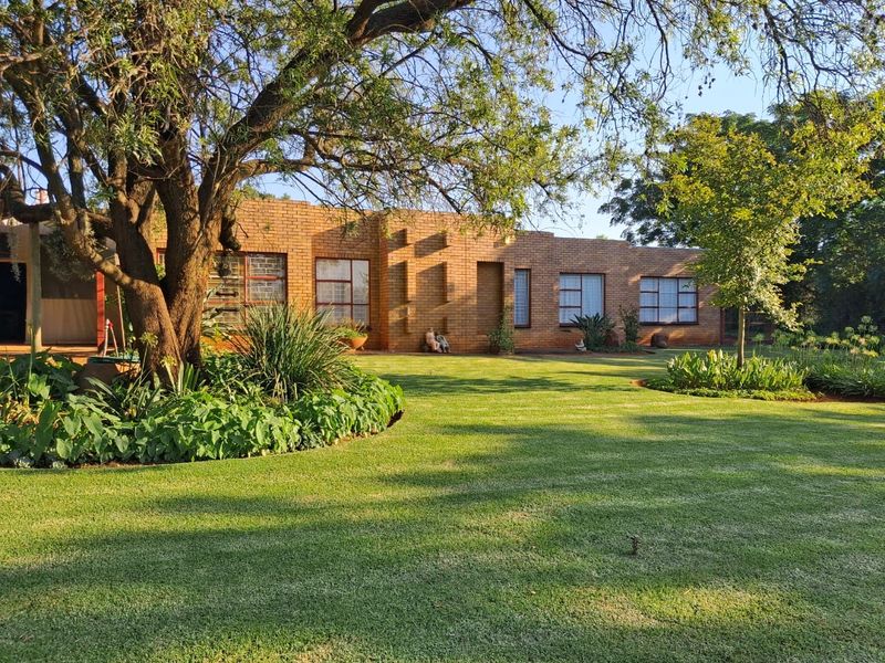 A well-maintained face brick farmhouse exterior featuring large windows, surrounded by a lush, manicured garden and mature trees.
