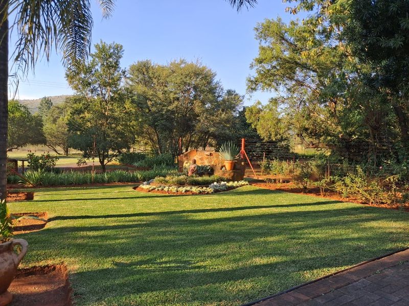 A well-maintained garden area featuring a stone feature wall, lush green lawn, and mature trees on a farm property.