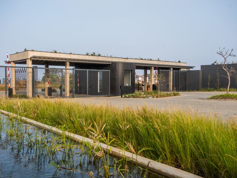 Modern security gatehouse entrance featuring a paved driveway, boom gates, and a landscaped water feature in a residential estate setting.