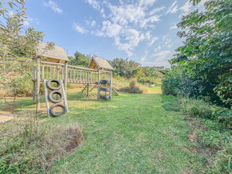 A spacious residential garden featuring a wooden playground structure set within a well-maintained lawn area surrounded by mature trees and lush greenery.