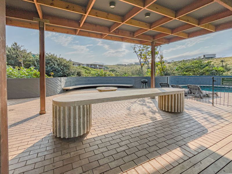 A spacious covered patio featuring a timber pergola, built-in concrete seating, and paved flooring, overlooking a private swimming pool and landscaped surroundings.