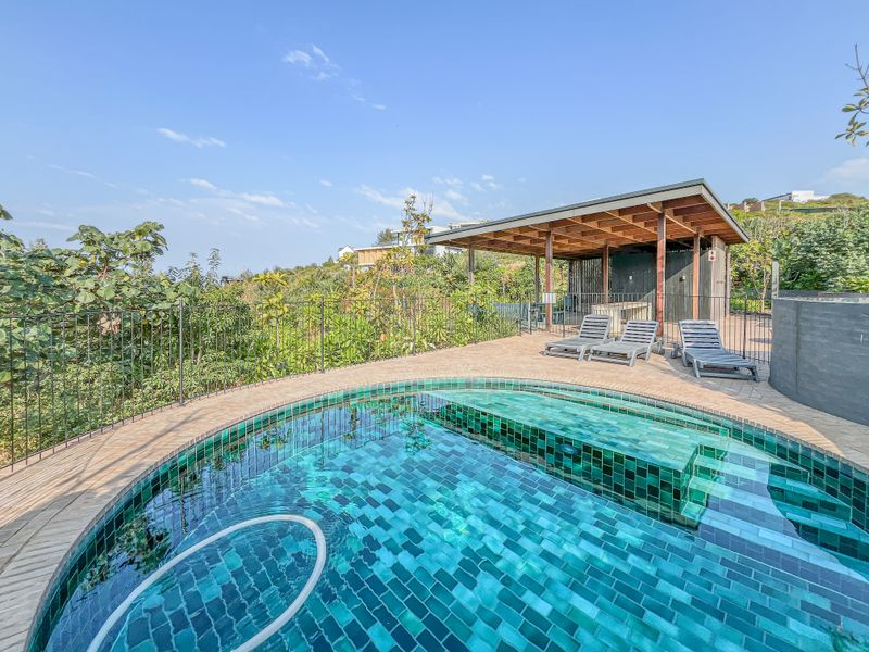 A well-maintained outdoor pool area featuring a tiled swimming pool, brick paving, and a covered patio structure set within a landscaped environment.