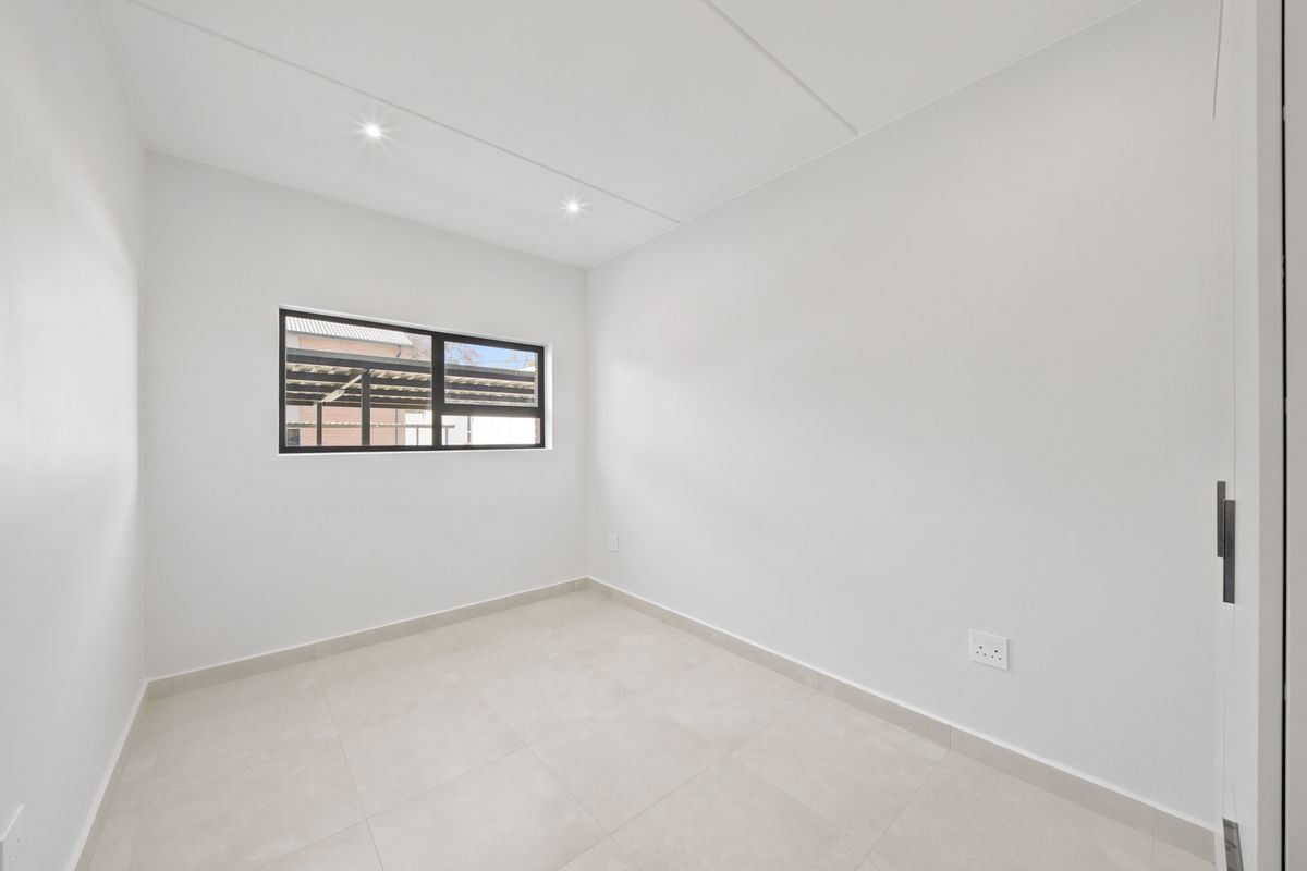 A bright, modern, and empty bedroom featuring neutral floor tiles, recessed ceiling lighting, and a black-framed window.