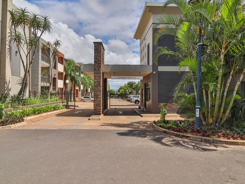 The secure entrance to Seasons Court apartment complex, featuring a gated access point, paved driveway, and landscaped garden beds.