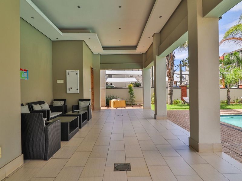 A spacious covered patio area featuring tiled flooring and recessed ceiling lighting, overlooking a communal swimming pool and landscaped garden.