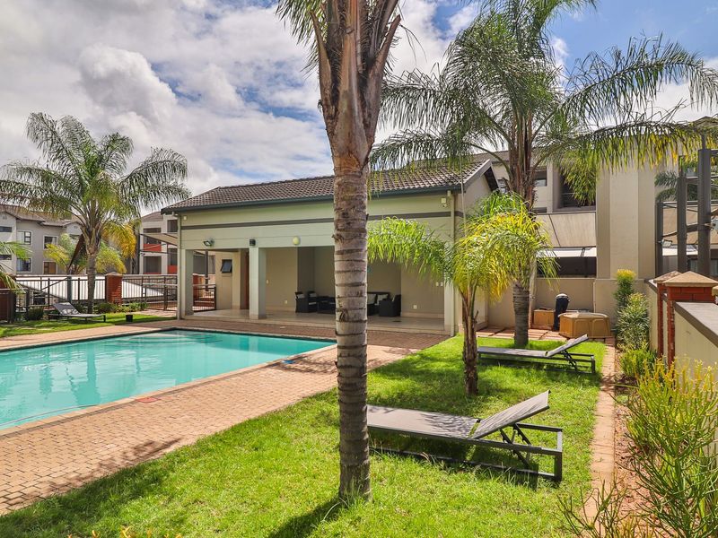 Communal swimming pool area featuring a paved deck, landscaped garden, and a covered outdoor entertainment pavilion within the apartment complex.