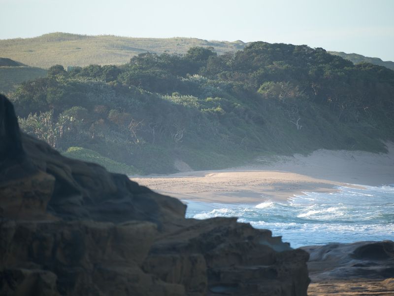 A scenic coastal view featuring natural rock formations, sandy beach, and lush green vegetation along the shoreline.