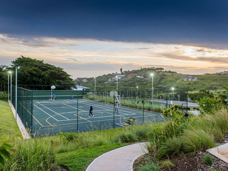 A well-maintained outdoor sports facility featuring a fenced tennis court with integrated basketball hoops and floodlighting, set within a landscaped residential estate.