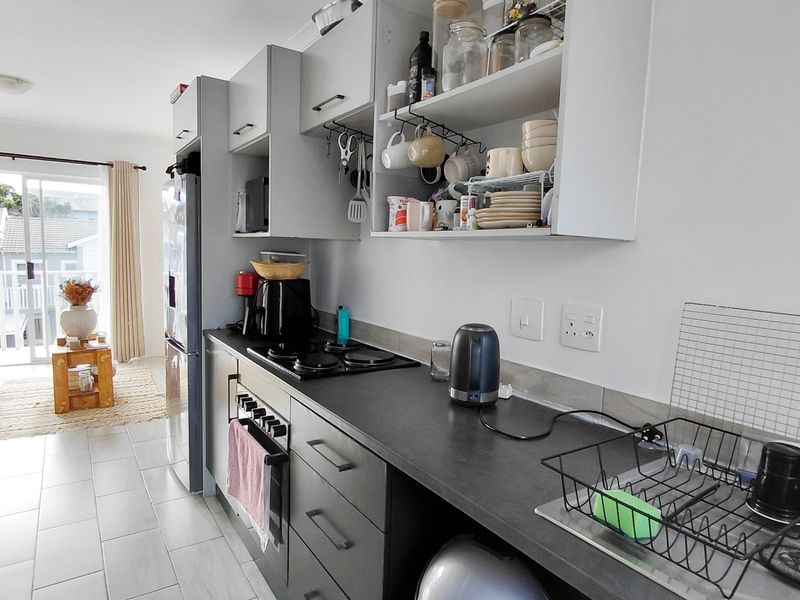 A functional galley-style kitchen featuring dark cabinetry, contrasting countertops, an integrated hob and oven, and tiled flooring, leading to an adjacent living area.