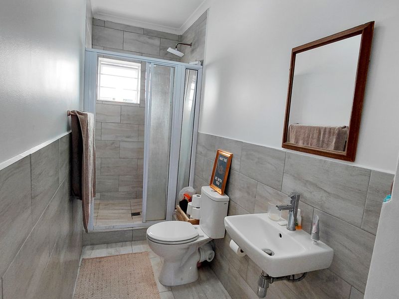 A modern bathroom featuring a tiled shower enclosure, wall-mounted basin with mixer tap, and a standard toilet with neutral grey wall and floor tiling.