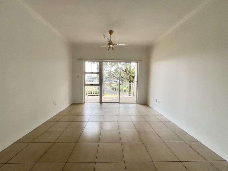 An empty, well-lit living room featuring neutral tiled flooring, a ceiling fan, and sliding glass doors leading to an outdoor balcony area.