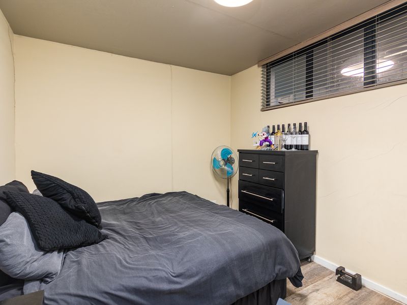 A compact bedroom featuring laminate flooring and a window with venetian blinds, showing signs of wear on the wall surfaces.