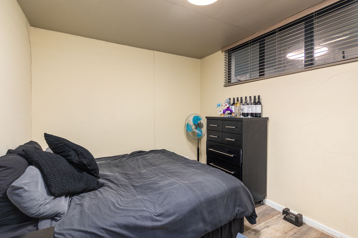 A compact bedroom featuring laminate flooring and a window with venetian blinds, showing signs of wear on the wall surfaces.