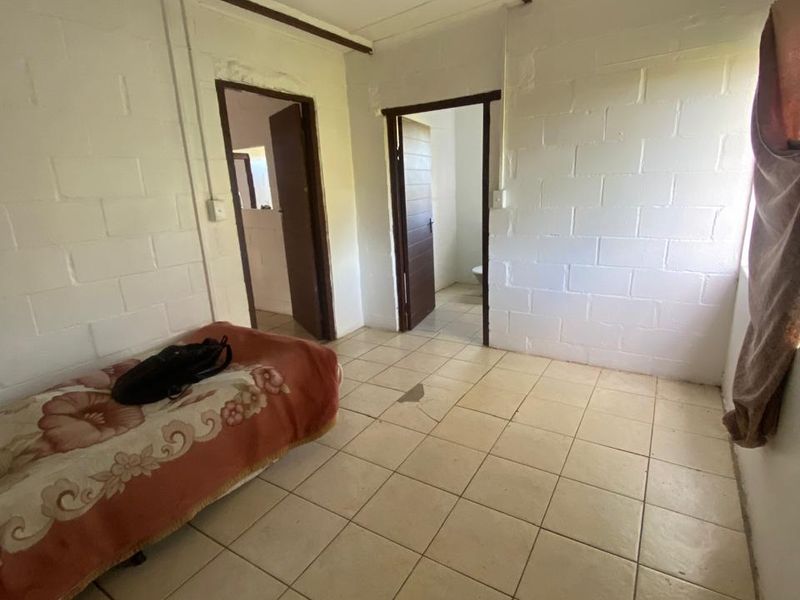 A simple bedroom featuring exposed blockwork walls, tiled flooring, and exposed ceiling beams, with direct access to an adjoining bathroom.