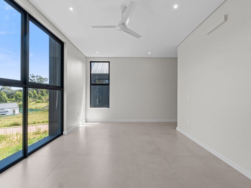 A bright, modern, and empty living area featuring large black-framed windows, tiled flooring, and a ceiling fan, offering a clean and minimalist aesthetic.