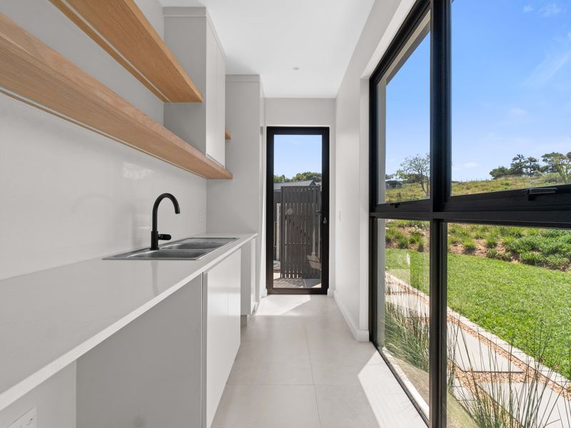 Modern scullery featuring white cabinetry, stone benchtops, floating timber shelves, and a large window overlooking the garden with direct outdoor access.