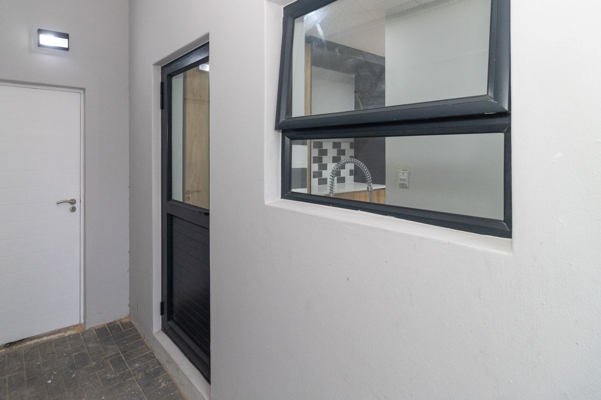 A clean, paved exterior rear area featuring modern aluminium framed doors and windows, with a view into the kitchen interior.