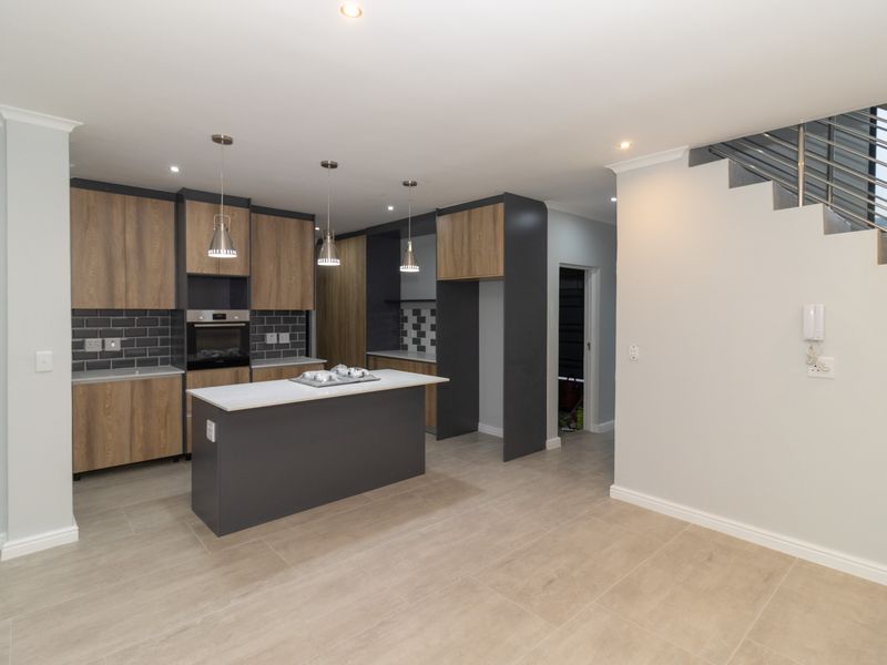 Modern open-plan kitchen featuring wood-tone cabinetry, stone countertops, a central island, built-in oven, and stylish subway tile backsplash, adjacent to a staircase.