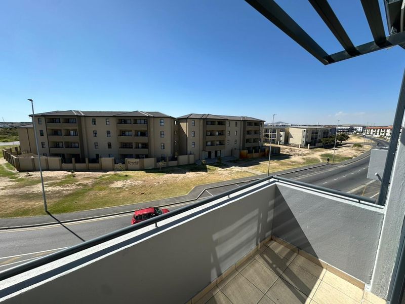 A private balcony featuring tiled flooring, a metal balustrade, and an overhead pergola, offering an elevated view of the surrounding apartment complex and street.