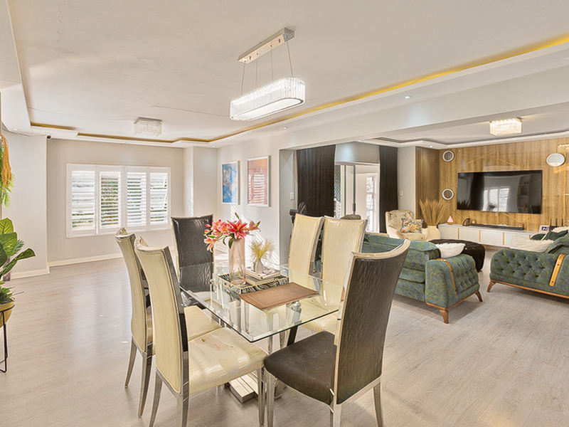 Spacious open-plan living and dining area featuring modern laminate flooring, recessed ceiling lighting, plantation shutters, and a decorative wood-paneled feature wall.