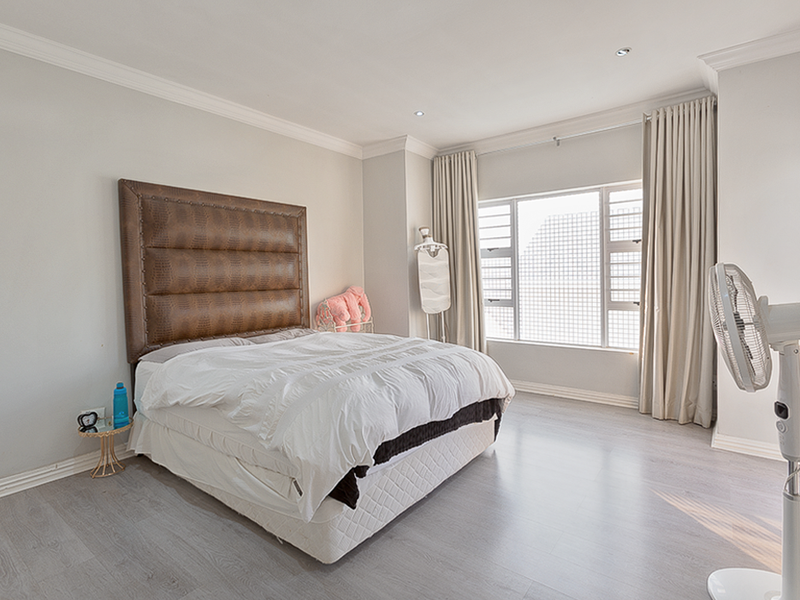 A well-maintained bedroom featuring light-toned laminate flooring, recessed ceiling lighting, decorative cornicing, and large windows with neutral curtains.