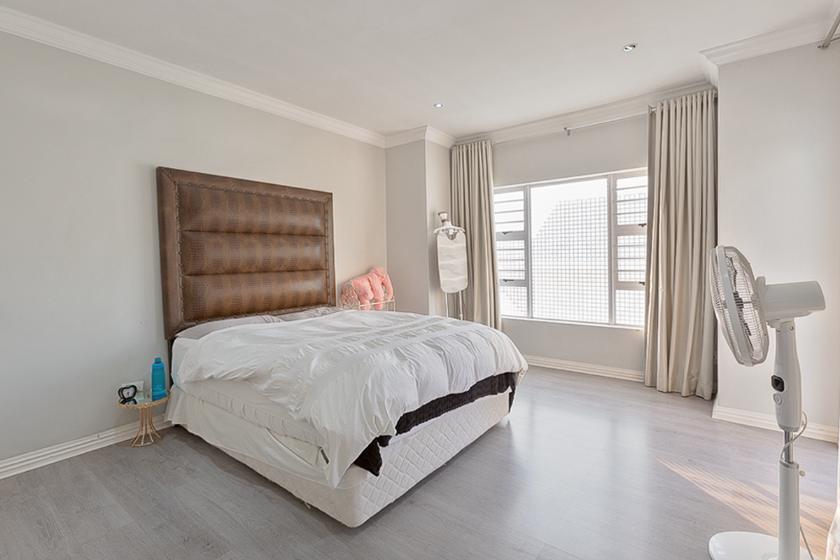 A well-maintained bedroom featuring light-toned laminate flooring, recessed ceiling lighting, decorative cornicing, and large windows with neutral curtains.