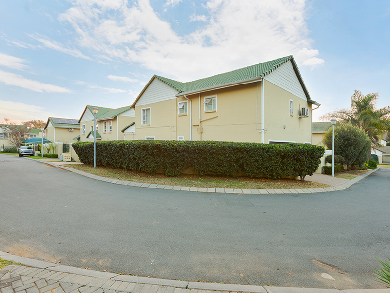 A view of the front exterior of a residential townhouse complex featuring a tiled roof, paved driveway, and established garden hedging.