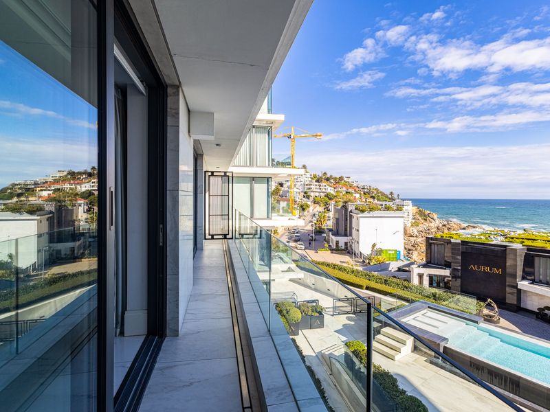 A modern, spacious balcony featuring high-quality marble-look tiling, a sleek glass balustrade, and expansive views of the coastline and surrounding luxury estate.