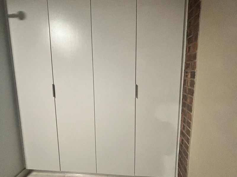 A clean bedroom space featuring modern white built-in cupboards, neutral tiled flooring, and an exposed brick accent wall.