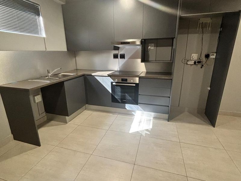 Modern kitchen featuring dark grey cabinetry, integrated oven and hob, stainless steel sink, and neutral floor tiles in an open-plan layout.