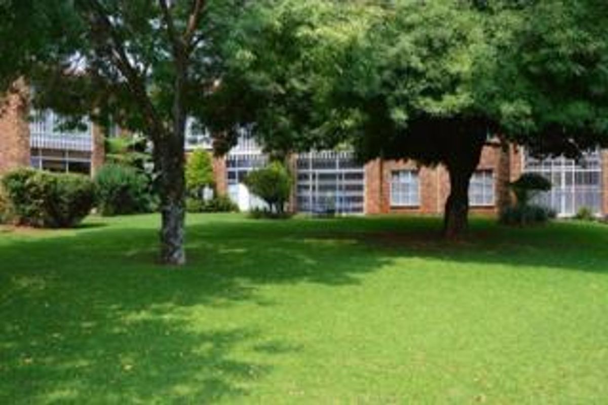 A view of the rear exterior of the townhouse complex, featuring a well-maintained lawn, mature trees, and brick facade with large windows.