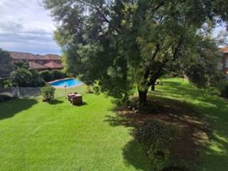 A spacious communal garden area featuring a well-maintained lawn, mature trees, and a swimming pool within the townhouse complex.