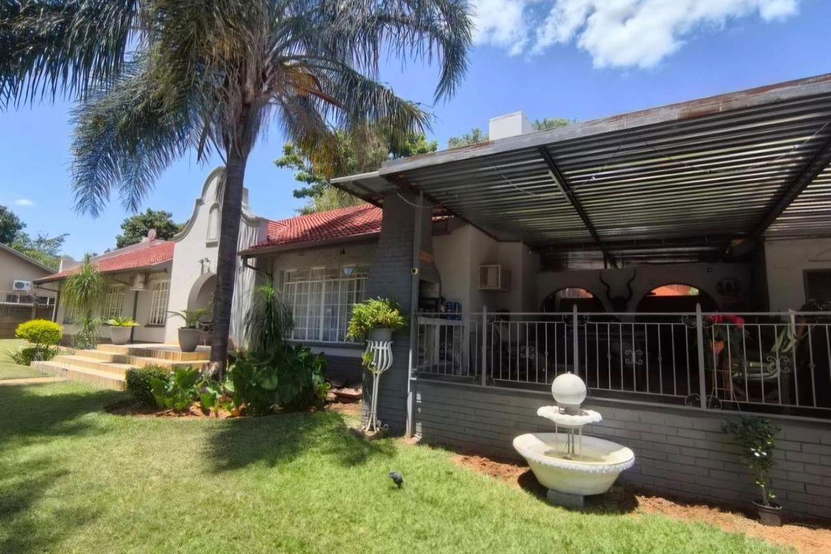 Rear view of the house featuring a covered patio with a built-in braai, metal fencing, and a landscaped garden with a stone fountain.