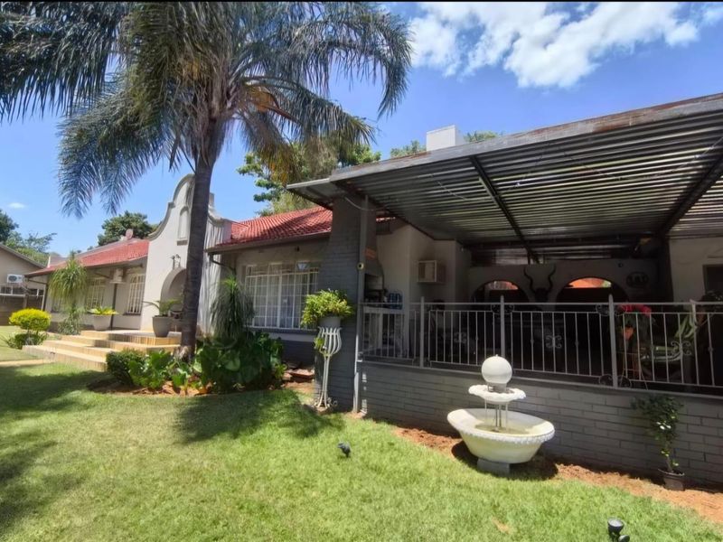Rear view of the house featuring a covered patio with a built-in braai, metal fencing, and a landscaped garden with a stone fountain.