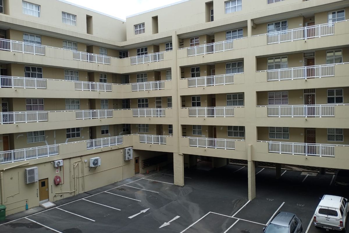 Exterior view of a multi-story apartment complex featuring private balconies, white railings, and a ground-level paved parking area.