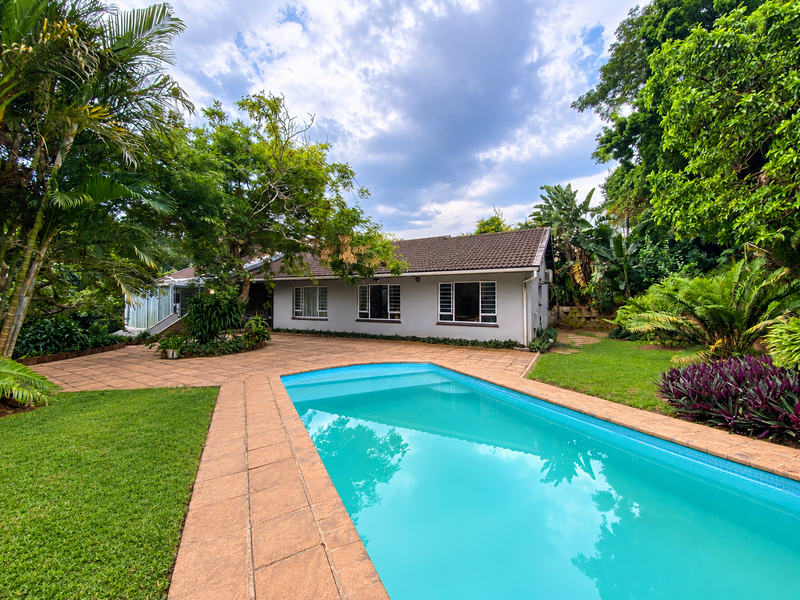 A well-maintained rear garden featuring a swimming pool with a tiled surround, established landscaping, and a clear view of the home's rear elevation.