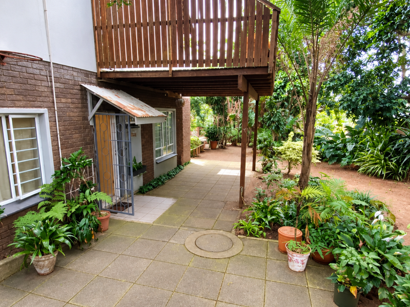 Rear exterior view featuring a paved patio area, a wooden balcony overhead, and a brick facade with a metal security gate entrance.