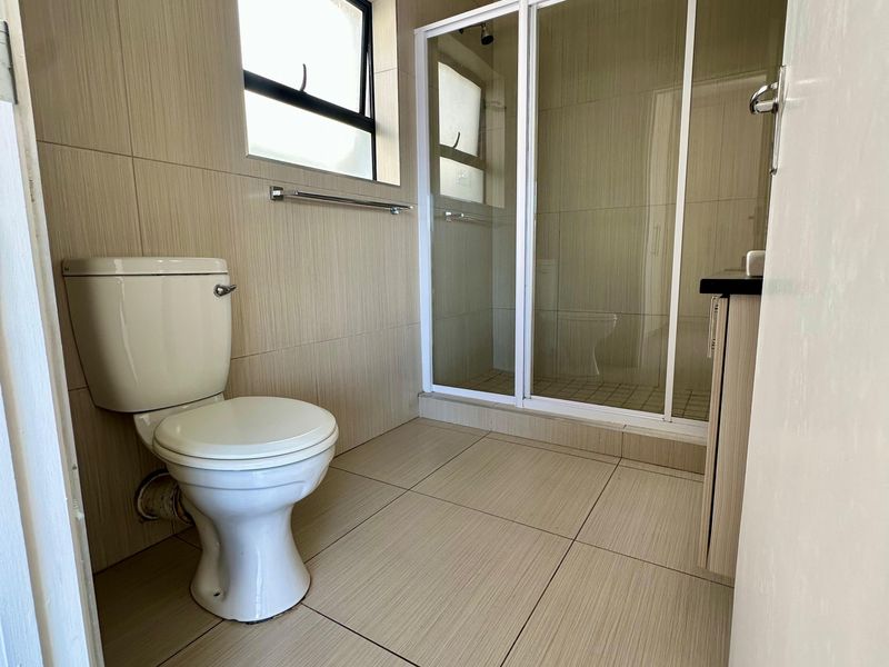 A clean, neutral-toned bathroom featuring a walk-in shower enclosure, a standard toilet, and a vanity cabinet with tiled finishes throughout.