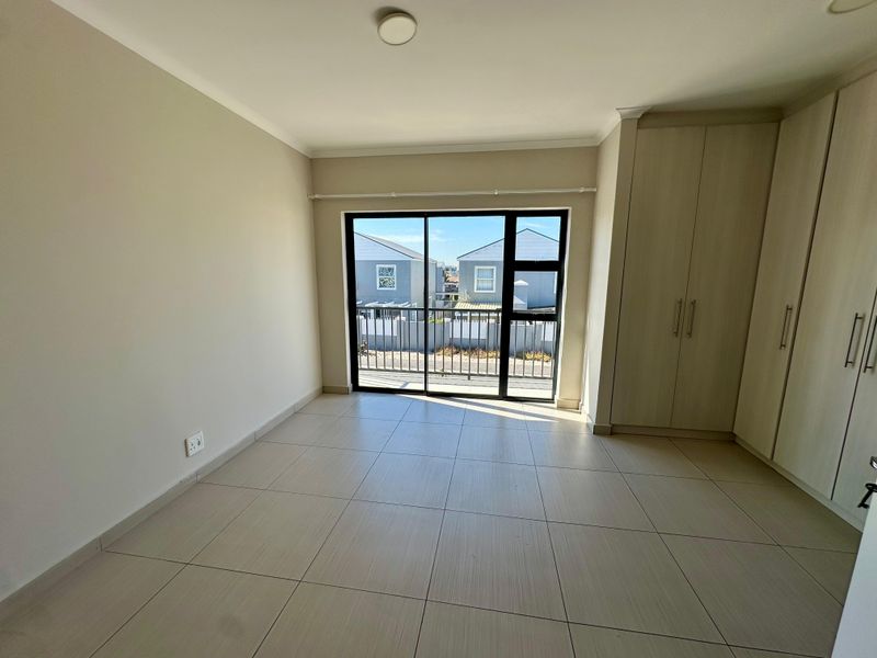A bright, empty bedroom featuring neutral tiled flooring, built-in cupboards, and a sliding glass door leading to a small balcony.